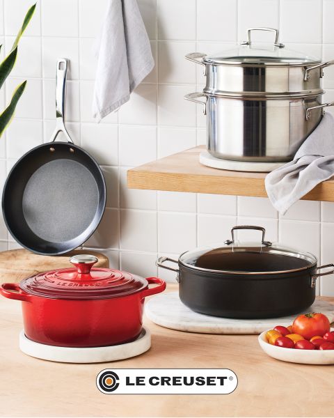 Une scène de cuisine avec une cocotte rouge, une poêle à frire antiadhésive, un ensemble de casseroles empilables en acier inoxydable, une marmite noire et un bol de tomates cerises sur un comptoir en bois clair devant un dosseret en carreaux blancs. Le logo Le Creuset est visible.
