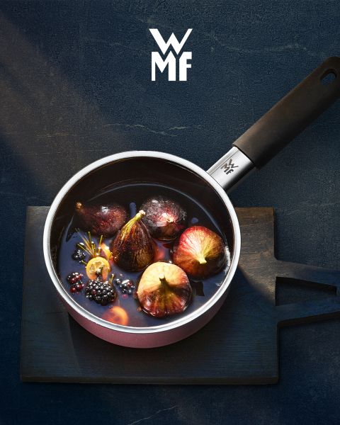 Une cocotte remplie de figues, de mûres et de garnitures aromatiques est posée sur une planche de bois sombre. Au-dessus de la cocotte, on aperçoit le logo WMF. L'arrière-plan est une surface sombre texturée.