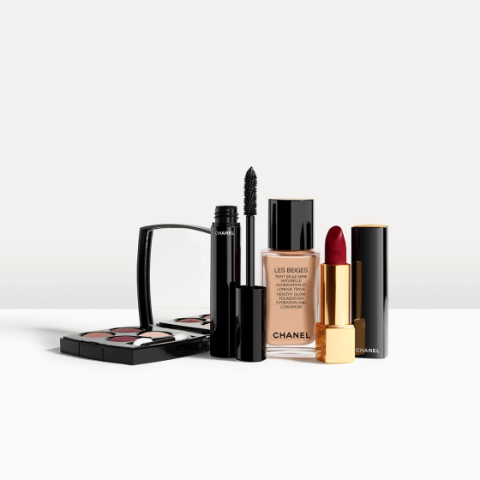 Une collection de produits de maquillage Chanel, comprenant un ensemble compact de fards à paupières, un mascara, un fond de teint liquide dans un flacon en verre et un rouge à lèvres rouge foncé dans un étui noir et or, disposés sur une surface blanche.