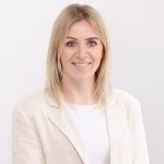 Une femme aux longs cheveux blonds sourit à la caméra. Elle porte un blazer clair sur un haut blanc. L'arrière-plan est simplement blanc, ce qui donne une apparence propre et professionnelle.