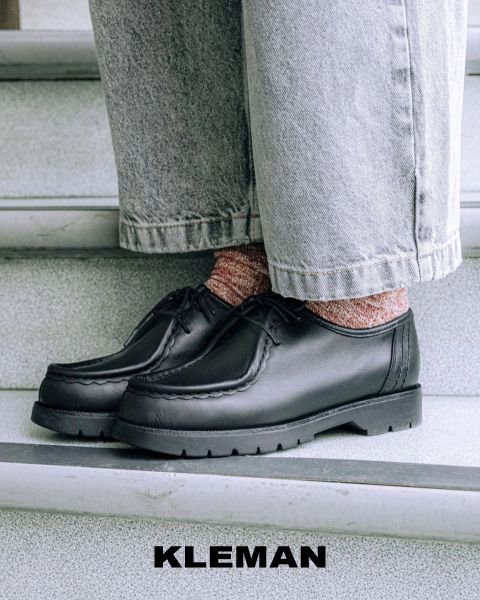 Gros plan d'une personne portant des chaussures en cuir noir avec des chaussettes à motifs roses et un jean ample et clair, debout sur un escalier gris. La marque "KLEMAN" apparaît en bas de l'image.