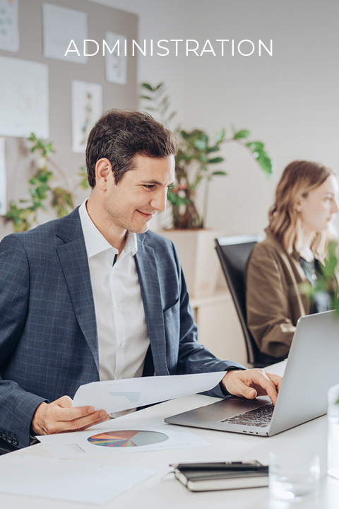 Un homme en costume travaille à un bureau avec un ordinateur portable et tient des papiers à la main. À l'arrière-plan, une femme est assise et travaille également. En haut, on peut lire le mot « Administration ». On peut voir des plantes et des diagrammes dans le bureau.