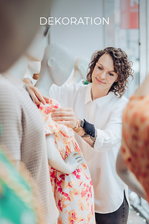 Une femme aux cheveux bouclés, portant des lunettes et une chemise blanche, ajuste une robe colorée sur un mannequin. La scène semble se dérouler dans un magasin de vêtements avec d'autres mannequins. En haut de l'image, on peut lire le mot « DÉCORATION ».