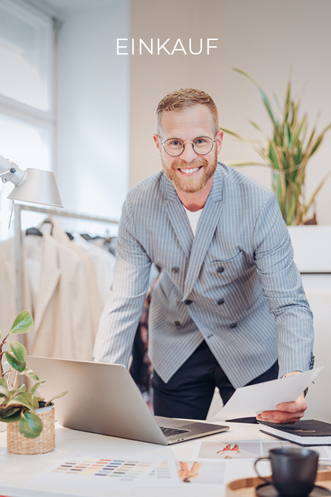 Homme souriant avec des lunettes et une barbe, penché sur un bureau, tenant des papiers à la main. Il porte un blazer rayé et se trouve dans un bureau moderne avec une plante et un ordinateur portable. Un porte-vêtements et des échantillons de couleurs sont visibles. Au-dessus de lui, on peut lire le mot « ACHATS ».