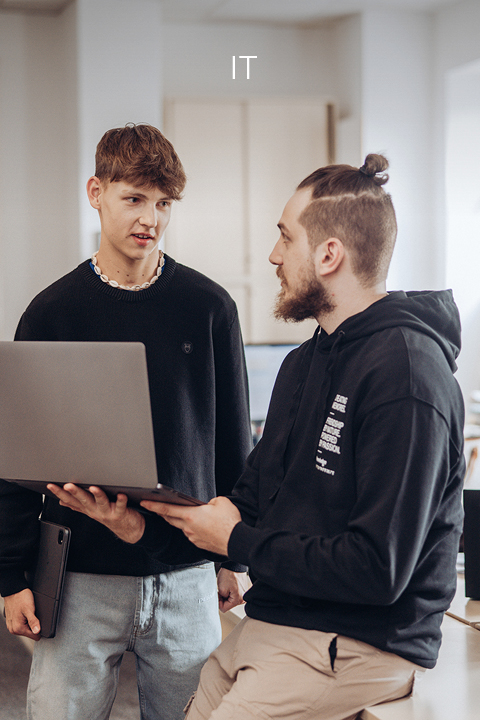 Deux hommes se tiennent dans un bureau. L'un est debout et tient un ordinateur portable à la main, l'autre est assis et discute. Les deux sont vêtus de manière décontractée. Le mot « IT » apparaît en haut de l'image et indique un contexte informatique.