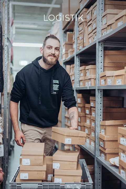 Un homme avec une barbe et un sweat à capuche noir range des cartons sur des étagères métalliques dans un entrepôt. Au-dessus de lui, on peut lire le mot « LOGISTIKI ». Les étagères sont remplies de cartons similaires.