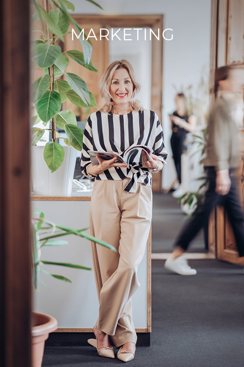 Une personne en chemise rayée et pantalon beige se tient dans un bureau, tenant un carnet ouvert à la main et souriant. Au-dessus, on peut lire le mot « Marketing ». À l'arrière-plan, une personne passe. Des plantes vertes luxuriantes décorent la pièce.