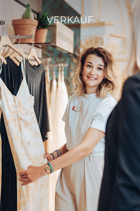 Une vendeuse aux cheveux blonds mi-longs sourit et tient une robe à fleurs dans un magasin de vêtements. Au-dessus, une pancarte indique « VENTE ». Le magasin a une lumière chaude et des plantes en pot.