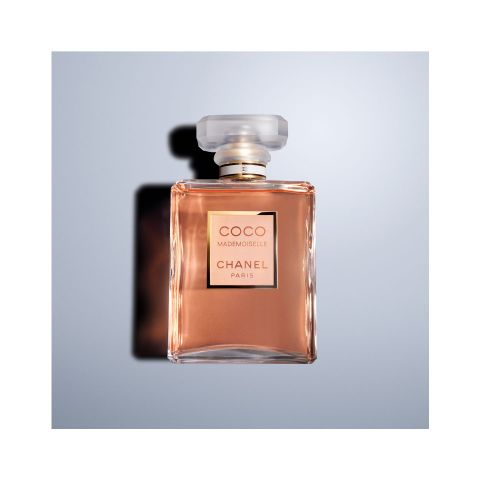 Un flacon carré en verre de parfum Coco Mademoiselle Chanel Paris contenant un liquide brun doré. Le flacon a un bouchon transparent à facettes et un col argenté. Il est photographié devant un fond bleu clair.
