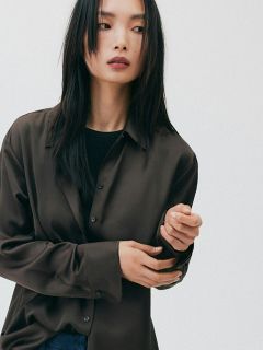 Femme aux longs cheveux noirs, vêtue d'une chemise marron ample et d'un haut noir, regardant sur le côté devant un simple fond blanc.