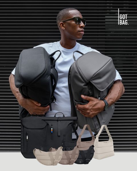 Un homme portant des lunettes de soleil tient de grands sacs gris devant un fond à rayures foncées. En dessous de lui, différents sacs de différentes tailles et couleurs sont soigneusement exposés. Le logo en haut à droite est « GOT BAG ».