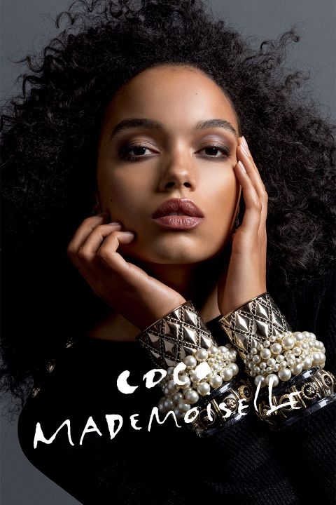 Une femme aux cheveux bouclés pose avec les mains devant le visage, présentant des bijoux élaborés et une tenue noire. Le texte « COCO MADEMOISELLE » apparaît en blanc en bas de l'image.