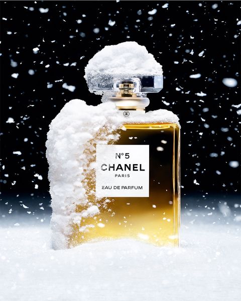 Un flacon de parfum Chanel N° 5 saupoudré de neige se dresse devant un fond sombre. Des flocons de neige tombent doucement autour du flacon, créant une scène hivernale. Le liquide doré dans le flacon transparent contraste avec la neige blanche.