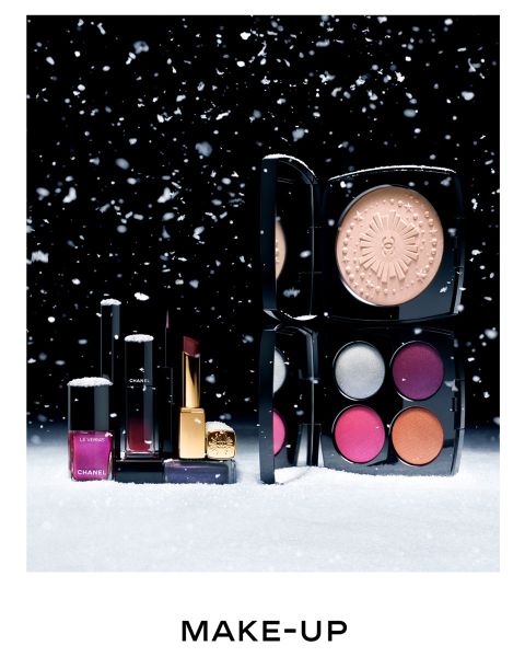 Une collection de produits de maquillage sur une surface enneigée avec un fond noir. Comprend un ensemble compact avec quatre ombres à paupières colorées, un illuminateur, des rouges à lèvres et du vernis à ongles. Des flocons de neige sont visibles dans l'air. En dessous, on peut lire le mot « MAKE-UP ».
