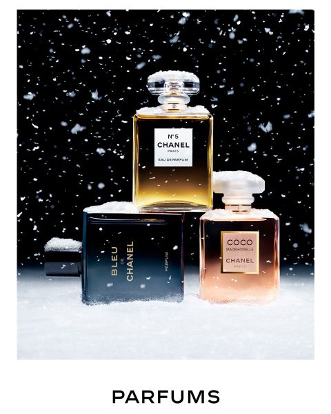Trois flacons de parfum Chanel, couverts de neige, sont disposés devant un fond noir avec des flocons de neige qui tombent. Les flacons portent les inscriptions N°5, BLEU et COCO MADEMOISELLE. En dessous, on peut lire le mot « PARFUMS ».
