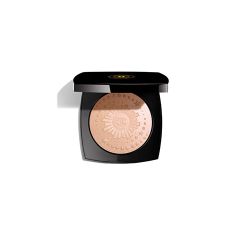 Un boîtier compact noir contenant une poudre pressée beige avec un motif complexe de rayons de soleil en relief sur sa surface. Le boîtier est ouvert, révélant la poudre. Un logo est visible sur le couvercle. Le fond est blanc.