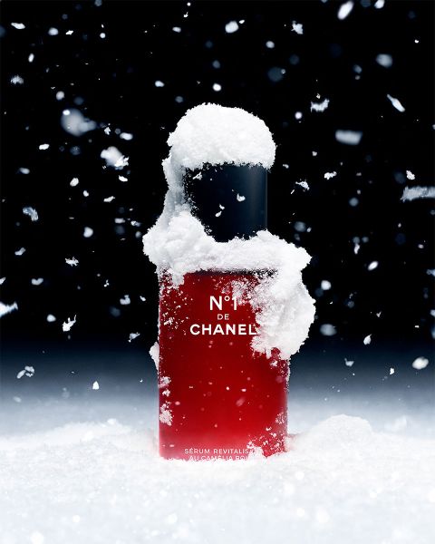 Un flacon rouge de sérum N°1 DE CHANEL, partiellement recouvert de neige, est posé verticalement devant un fond sombre. Des flocons de neige tombent, conférant au produit une atmosphère hivernale.