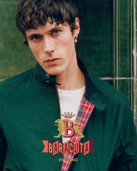 Une personne aux cheveux courts et foncés, portant une boucle d'oreille et une veste foncée sur une chemise blanche, regarde la caméra. L'intérieur de la veste est à carreaux rouges. Le logo Baracuta avec un lion et un "B" est visible sur le devant.