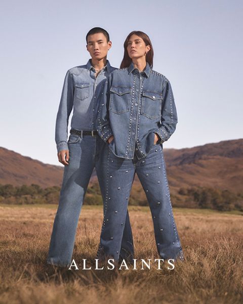 Deux personnes se tiennent dans un champ dégagé, vêtues de tenues en jean ornées de clous. La personne à gauche porte une chemise en jean claire et un jean, tandis que la personne à droite porte une veste en jean foncée et un jean. En bas, il est écrit « ALLSAINTS ».