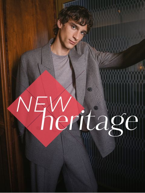 Un homme en costume gris se tient devant un mur en bois sur lequel est inscrit « NEW heritage » en gras.