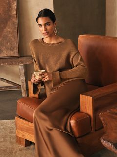 Une femme vêtue d'un pull marron et d'une longue jupe marron est assise sur un fauteuil en cuir et tient une tasse à la main ; à l'arrière-plan, un intérieur aux tons neutres est visible.