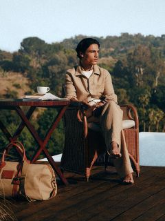 Une personne en tenue légère est assise pieds nus à une table avec du café, en plein air au bord de la piscine, avec des arbres et des collines en arrière-plan. À côté de la table se trouve un sac.