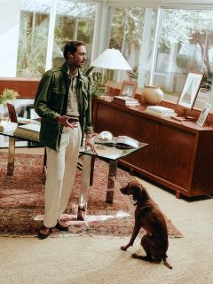 Un homme se tient debout avec un téléphone à la main à côté d'un bureau dans un bureau baigné de soleil, tandis qu'un chien marron est assis sur un tapis à motifs et lève les yeux vers lui.