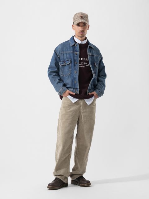 Un homme en veste en jean, pantalon en velours côtelé, casquette à carreaux, pull et chaussures marron se tient devant un simple fond blanc.