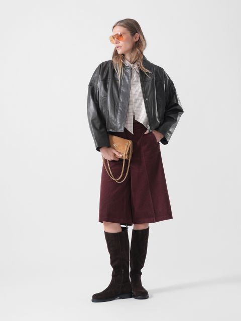 Une femme vêtue d'une veste en cuir noire, d'un pantalon culotte bordeaux, de bottes hauteur genou et de lunettes de soleil, tenant un sac à main marron, posant devant un simple fond blanc.