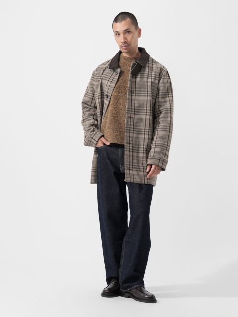 Un homme portant un manteau à carreaux, un pull marron, un jean foncé et des chaussures noires se tient devant un simple fond blanc.