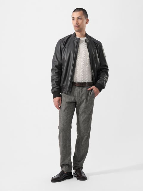 Un homme portant une veste en cuir noire, un pull blanc, un pantalon gris et des chaussures foncées, se tient devant un simple fond blanc et regarde légèrement sur le côté.