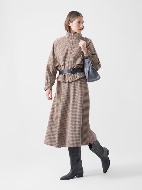 Une femme vêtue d'une jupe marron à motifs et d'une veste avec une ceinture noire, des bottes grises et un sac à main gris, posant devant un simple fond blanc.
