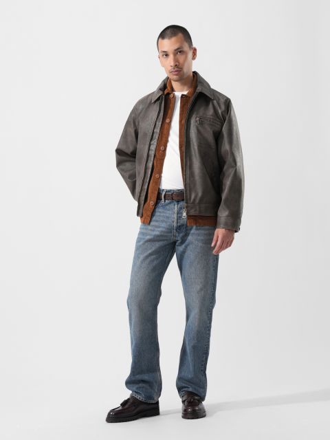 Sur un fond uni, un homme porte une veste marron sur une chemise, un jean bleu et des chaussures foncées.