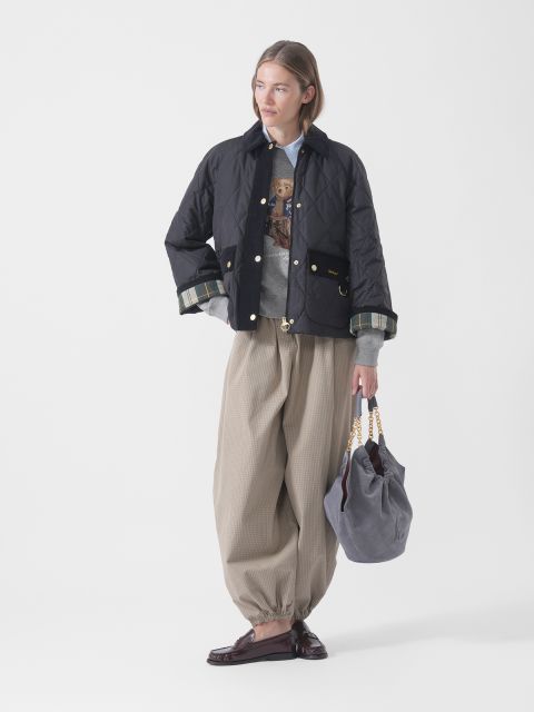 Une personne vêtue d'une doudoune noire, d'un pull gris, d'un pantalon beige ample et de mocassins tient un sac gris avec des poignées en chaîne dorées et se tient devant un fond blanc.