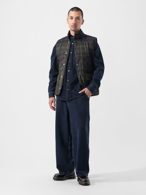 Sur un fond blanc uni, une personne porte un gilet matelassé foncé par-dessus une chemise boutonnée bleu marine et un pantalon large bleu marine, avec des chaussures foncées.
