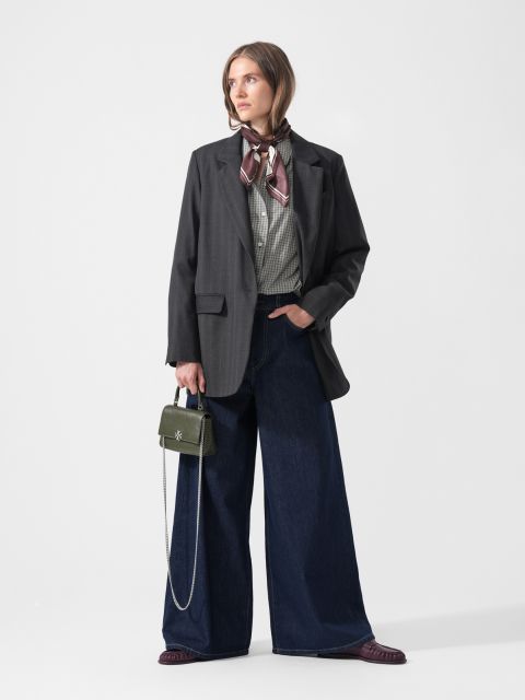 Une femme en jean large, chemise à carreaux, blazer foncé et écharpe, tenant un petit sac à main vert, se tient devant un simple fond blanc.