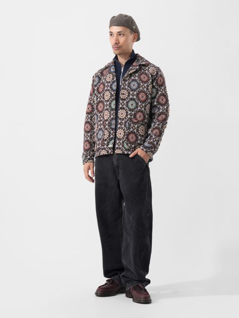Un homme en veste à motifs, pantalon noir, chaussures bordeaux et béret gris se tient devant un simple fond blanc.