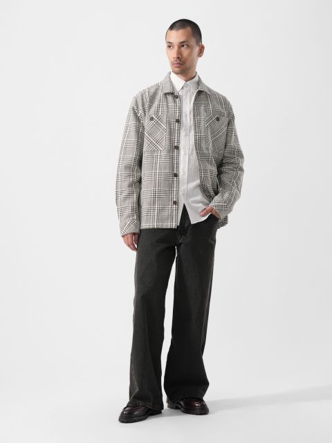 Sur un fond blanc uni, un homme porte une veste à carreaux sur une chemise blanche, un pantalon noir ample et des chaussures noires.