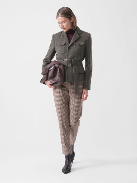 Une femme en blazer ceinturé, pantalon à carreaux et bottes tient un sac marron, regarde vers le bas et se tient devant un simple fond blanc.