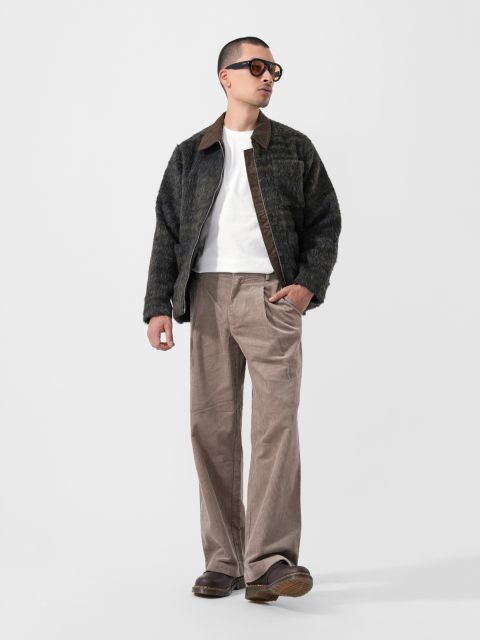 Un homme avec des lunettes de soleil, une veste foncée et moelleuse, un t-shirt blanc, un pantalon large beige et des chaussures marron se tient devant un simple fond blanc.