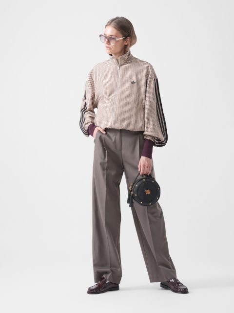Une personne portant des lunettes de soleil, une veste à carreaux, un pantalon gris ample et des chaussures bordeaux, tient un petit sac rond noir devant un fond blanc uni.
