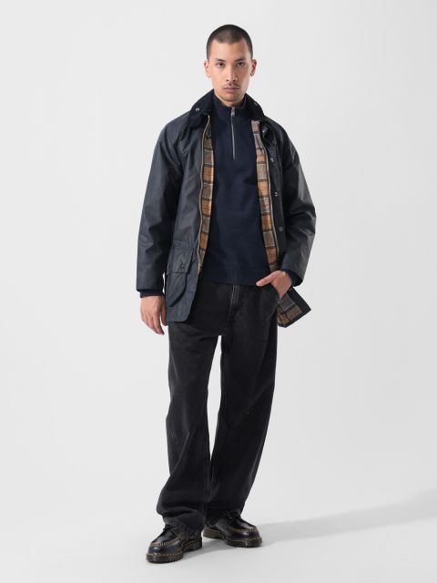 Un homme en tenue décontractée se tient devant un simple fond blanc : veste foncée avec doublure à carreaux, pull bleu marine zippé, jean noir ample et chaussures noires.