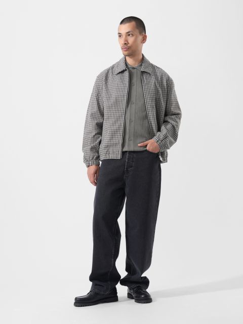 Sur un fond blanc uni, un homme porte une veste à carreaux grise, une chemise grise, un jean noir ample et des chaussures noires.