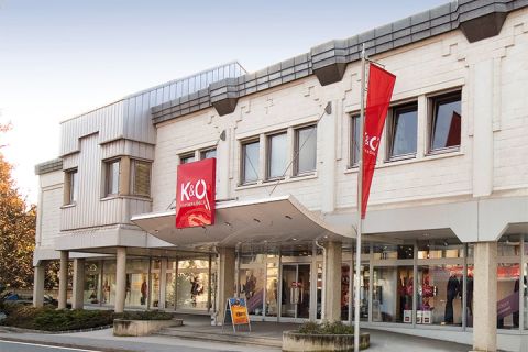 Le bâtiment moderne à la façade beige abrite les magasins K&Ö Kastner & Öhler et Giga X Sport. Des panneaux rouges et orange sont bien visibles. De grandes fenêtres et une rue devant, sous un ciel clair, complètent le tableau.
