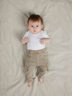 Un bébé souriant est allongé sur une couverture blanche, portant un pull en tricot bleu, un pantalon en velours côtelé marron avec des poches et des chaussettes grises. Le bébé a l'air heureux et détendu, avec les bras légèrement pliés au niveau des coudes.