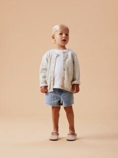 Une jeune fille souriante en survêtement lavande se tient devant un fond gris et porte un très grand nœud blanc sur la tête.