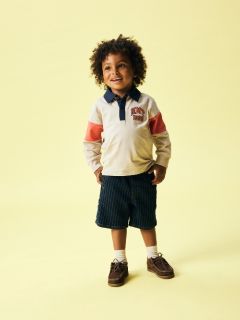Un garçon aux cheveux blonds porte un bonnet noir, une veste polaire crème sur une chemise rayée, un jean gris et des baskets. Il se tient devant un fond bleu uni et regarde vers le bas, les mains jointes.