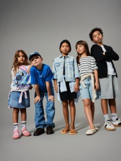 Une petite fille aux cheveux blonds se tient souriante devant un fond vert, portant un pull sans manches marron sur une robe à fleurs, des chaussettes montantes et des chaussures marron.
