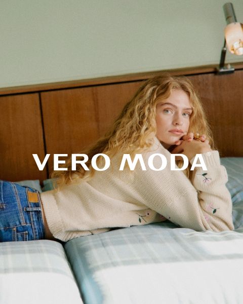 Une femme portant des lunettes et une robe à fleurs à volants est appuyée contre un mur blanc devant un fond carrelé bleu. L'inscription "VERO MODA" apparaît au centre de l'image.
