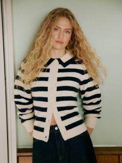 Une femme aux cheveux bouclés se tient devant un mur carrelé. Elle porte un cardigan gris clair duveteux, un short en cuir noir taille haute et des collants noirs, les mains dans les poches et une expression confiante sur le visage.